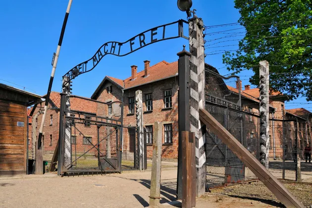 The 'Arbeit Macht Frei' sign over the entrance gate to a historical camp, with barbed wire fences and brick buildings under a clear blue sky.