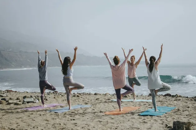 Un gruppo WeRoad di cinque donne in viaggio, che praticano yoga in posizione dell'albero su tappetini su una spiaggia sabbiosa di fronte all'oceano.