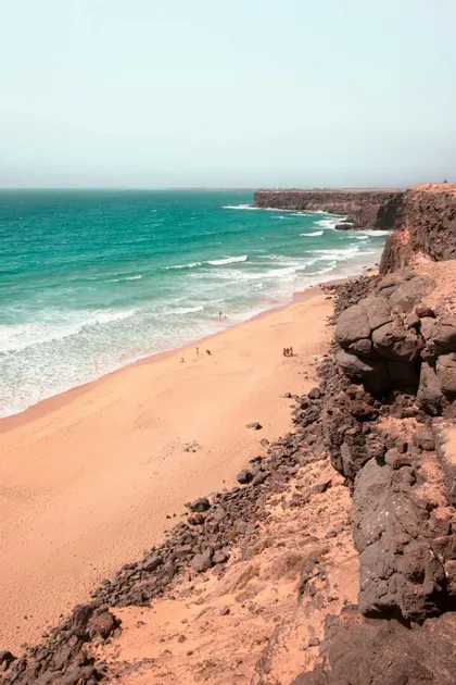 Una vista da una scogliera rocciosa che si affaccia su un'ampia spiaggia sabbiosa con acqua turchese e piccole onde che si frangono sulla riva.