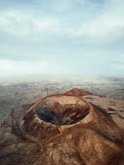 Vista aerea di un grande cratere vulcanico color ruggine in un vasto paesaggio arido con una città lontana sotto un cielo nuvoloso.