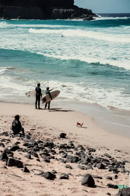 Due surfisti in muta tengono le loro tavole da surf sulla battigia di una spiaggia sabbiosa, ammirando le onde turchesi.
