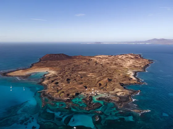 Una vista aerea di una piccola isola vulcanica arida circondata da acqua turchese e dal mare blu profondo, con barche a vela ancorate al largo.