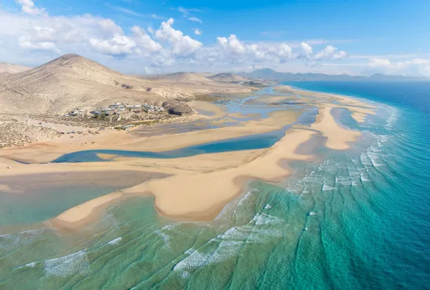 Vista aerea di una vasta costa sabbiosa che incontra le acque turchesi dell'oceano, con colline aride e un piccolo villaggio in lontananza.
