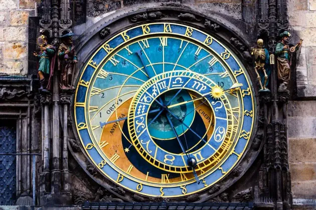 Un primer plano de un reloj astronómico ornamentado con esferas azules y doradas, signos del zodiaco y figuras talladas en un edificio de piedra.
