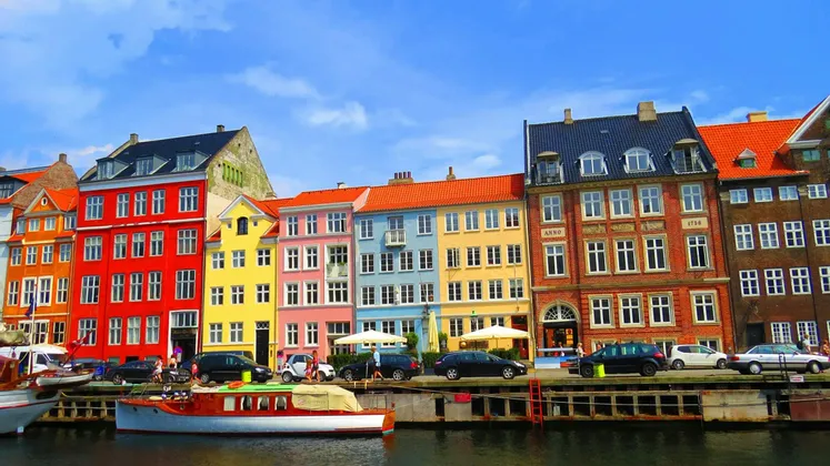 Une rangée de bâtiments aux couleurs vives longe un canal, avec des bateaux amarrés sur l'eau et des voitures garées le long de la rue.