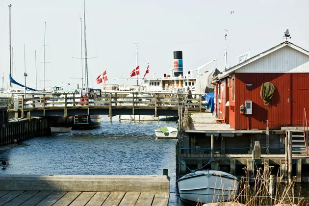 Des voiliers aux drapeaux danois amarrés dans un port ensoleillé, à côté d'un bâtiment en bois rouge et d'un pont piéton.