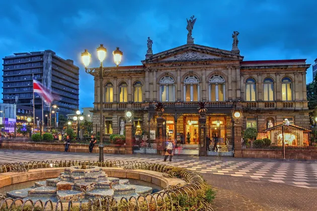 Il sontuoso edificio del Teatro Nazionale in Costa Rica, illuminato al crepuscolo, con una fontana decorativa in primo piano in una piazza cittadina.