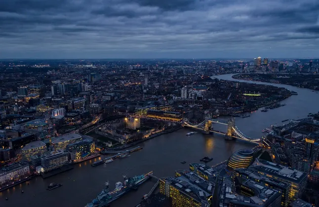 Una vista aerea del panorama urbano di Londra al tramonto, con il Tower Bridge illuminato e il Tamigi sotto un cielo nuvoloso.
