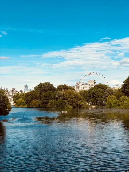 Una vista sul lago in un parco, con una grande ruota panoramica e lo skyline della città visibile dietro una fila di alberi verdi.