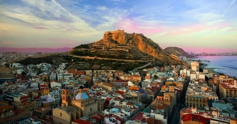Una veduta sopraelevata di una città costiera, con un'imponente fortezza su un colle, immersa nella luce di un tramonto dai colori vivaci.