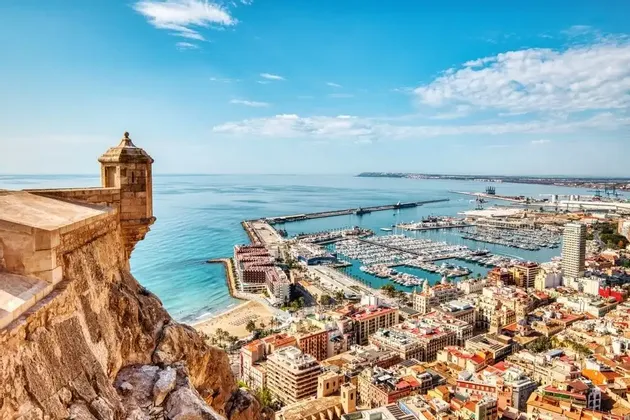 Veduta dall'alto da una fortificazione in pietra che si affaccia su una città costiera con un'ampia marina piena di barche sotto un cielo soleggiato.