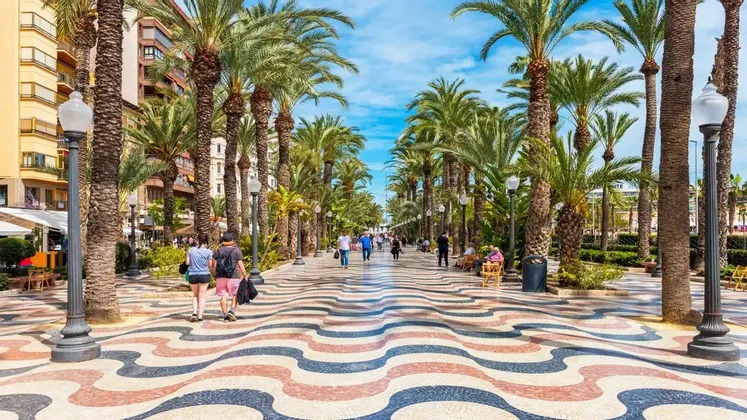 Le persone passeggiano lungo un ampio lungomare fiancheggiato da alte palme, caratterizzato da una pavimentazione a mosaico colorata e ondulata sotto un cielo azzurro.