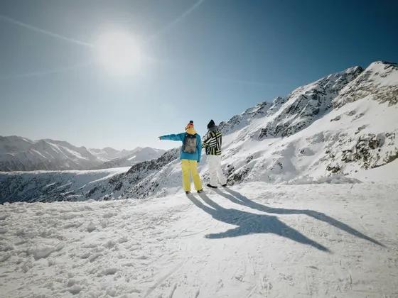 Deux personnes d'un voyage de groupe WeRoad se tiennent sur un sommet de montagne enneigé, pointant la vue sous un soleil éclatant.