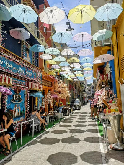 Sombrillas de colores pastel cuelgan suspendidas sobre una calle estrecha con cafés al aire libre, proyectando sombras distintas sobre el pavimento.