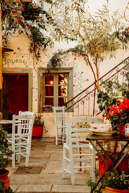 Un patio de cafetería al aire libre con sillas blancas ubicadas en un patio de piedra, junto a un edificio con enredaderas pintadas y plantas en macetas.