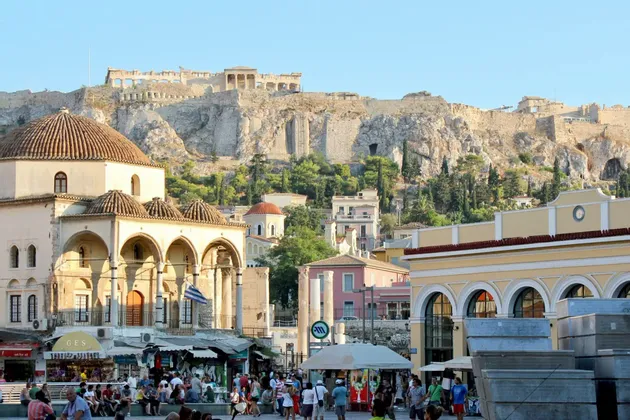Ruinas antiguas se alzan sobre una gran colina rocosa, dominando una bulliciosa plaza de la ciudad con edificios históricos abovedados y multitudes de gente.