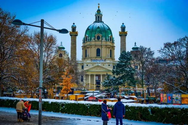 Una grande chiesa con una cupola verde domina un parco innevato dove le persone passeggiano per un mercatino invernale.