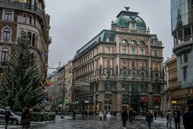 I pedoni passeggiano in una piazza cittadina innevata, con un grande albero di Natale e sontuosi edifici storici, sotto un cielo coperto.
