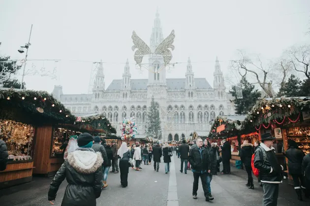Una folla di persone passeggia in un mercatino di Natale all'aperto, con bancarelle decorate, di fronte a un grande edificio storico.