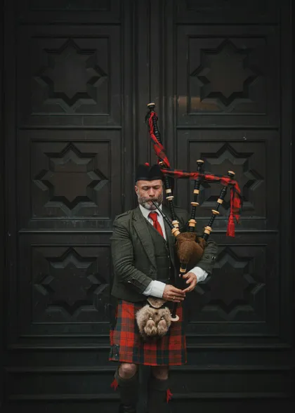 Un uomo in abito tradizionale scozzese, inclusi kilt e giacca di tweed, tiene una cornamusa mentre si trova di fronte a una grande porta di legno intarsiata.