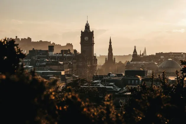 Un paesaggio urbano storico con una prominente torre dell'orologio e guglie, stagliate contro un'alba calda e velata, visto da dietro le cime scure degli alberi.