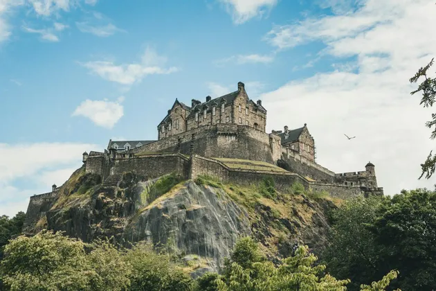 Un castello storico in pietra si erge su una scogliera rocciosa sopra alberi verdi, sotto un cielo azzurro parzialmente nuvoloso.