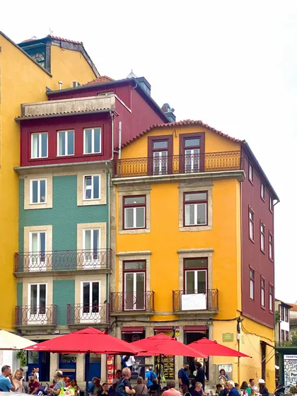 Edificios coloridos en amarillo, verde azulado y granate se alzan sobre un café callejero donde la gente se sienta bajo sombrillas rojas.