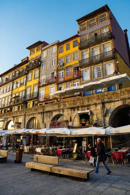 Edificios coloridos y con azulejos dan a un paseo de piedra con cafeterías al aire libre y bancos bajo un cielo despejado durante la hora dorada.