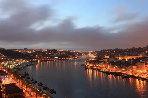 Un horizonte urbano iluminado a lo largo de un ancho río al anochecer, con las luces de los edificios reflejándose en el agua y un puente a lo lejos.