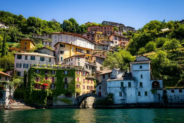 Colorful buildings of a village are built into a lush green hillside that meets the water, with a small stone bridge in the foreground.