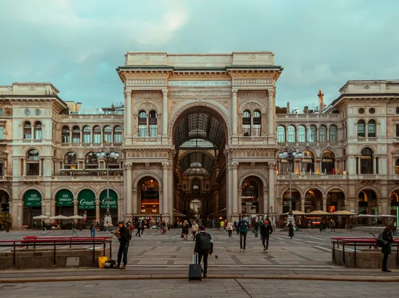 People walk across a large paved plaza in front of a grand, ornate building with a central archway under an overcast sky.