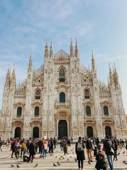 The ornate, spired facade of a large gothic cathedral with a crowd of people and pigeons gathered in the square below.
