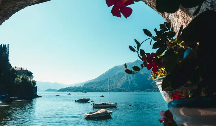 A view of a large lake with several boats, seen from under a stone archway with pink flowers in the foreground and mountains in the background.