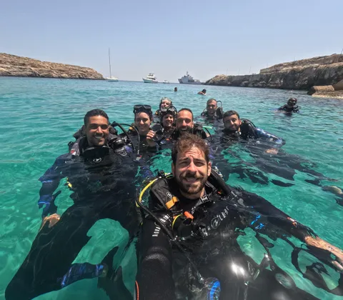 Un gruppo WeRoad in gita subacquea sorride per un selfie mentre galleggia in acque limpide e turchesi vicino a una costa rocciosa.