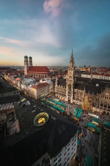 Una vista aerea di una piazza cittadina gremita di mercatini di Natale, circondata da edifici storici e un grande albero di Natale al tramonto.