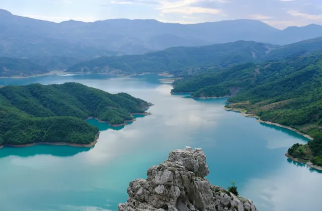 Da un punto panoramico roccioso, un lago turchese brillante si snoda tra verdi colline, con catene montuose in lontananza.