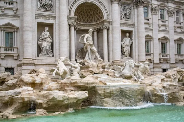 Una ornamentada fuente de mármol con grandes esculturas de figuras y caballos, con agua fluyendo sobre rocas hacia una piscina turquesa.