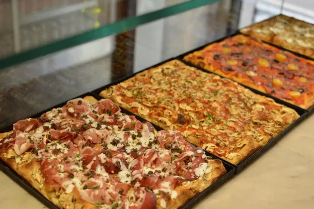 Una hilera de pizzas rectangulares con varios ingredientes, como jamón serrano y champiñones, expuestas en bandejas negras detrás de un mostrador de cristal.