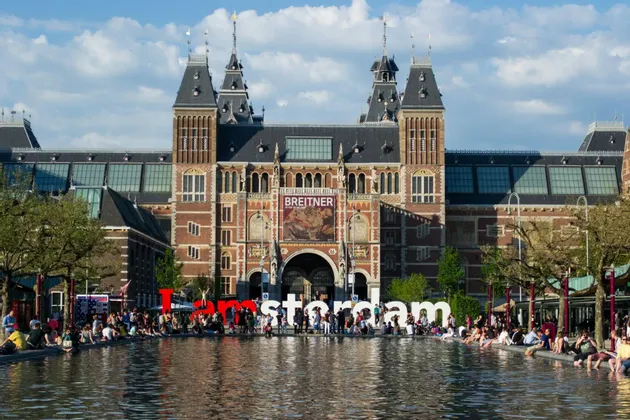 L'iconica insegna 'I amsterdam' di fronte al Rijksmuseum, con persone sedute attorno al bordo di una grande vasca riflettente.