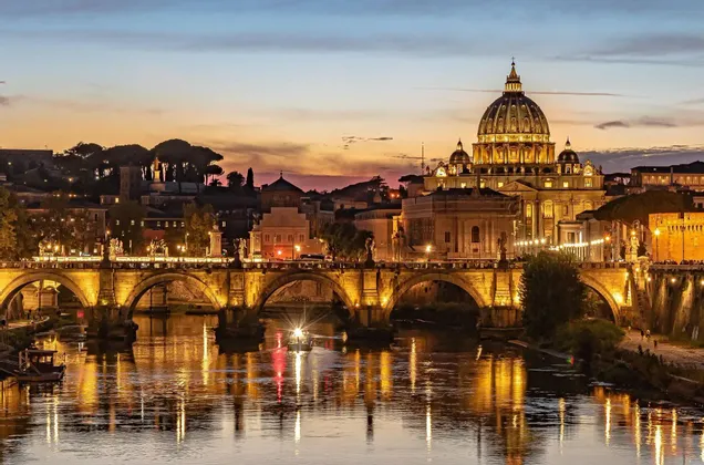 Una gran basílica iluminada y un puente de piedra arqueado se reflejan en un río durante un atardecer colorido.