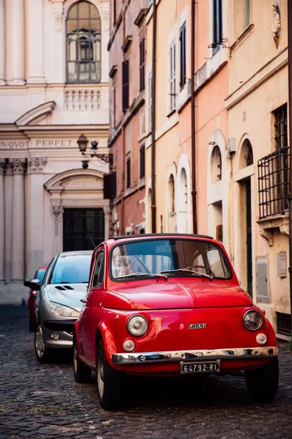 Un Fiat 500 rojo vintage aparcado en una estrecha calle empedrada con edificios históricos de fondo.