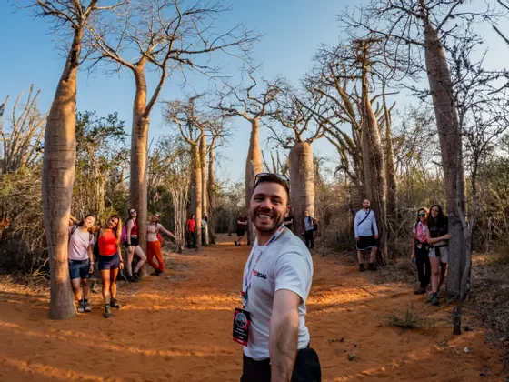 Un uomo sorridente si scatta un selfie con il suo gruppo WeRoad tra grandi baobab su un sentiero sterrato.
