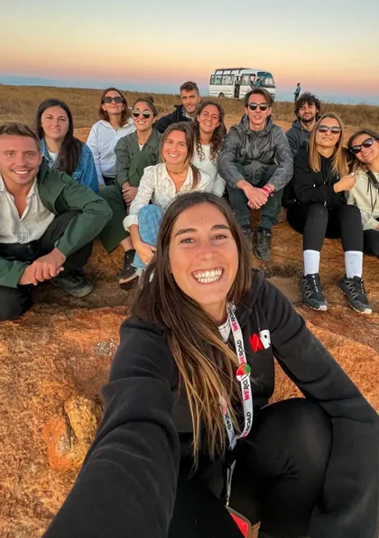 Una donna si scatta un selfie con il suo gruppo di viaggio WeRoad mentre sono seduti su uno sperone roccioso al tramonto.