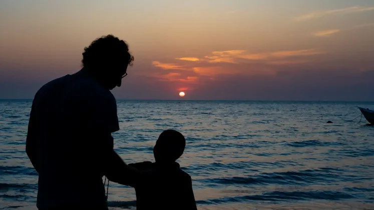 Una silhouette di un adulto e un bambino che ammirano il tramonto sull'oceano dalla spiaggia.