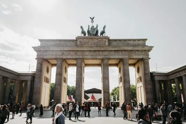 Un arco di trionfo neoclassico sormontato da una quadriga, ammirato da una piazza vivace e affollata sotto il sole.