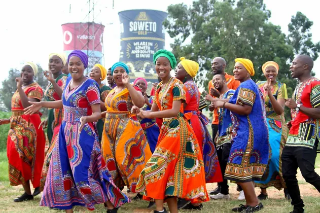 Un groupe de personnes vêtues de tenues africaines traditionnelles et colorées, dansant et chantant ensemble en plein air sur un terrain herbeux.