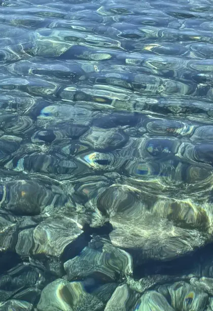 La luce del sole si riflette sulla superficie dell'acqua limpida e increspata, rivelando sul fondo pietre lisce e scure.
