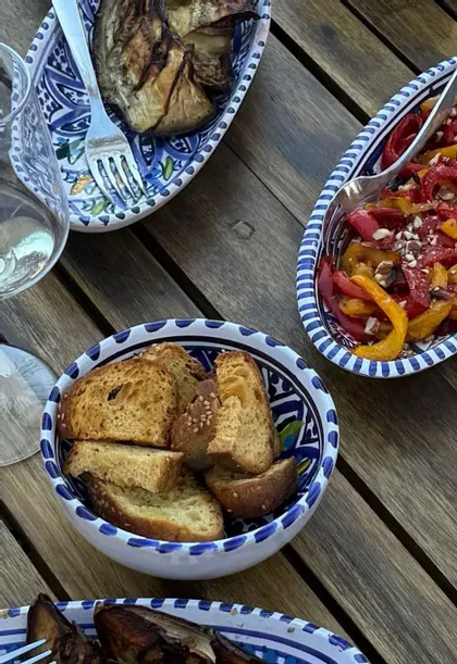 Una vista dall'alto di antipasti mediterranei, tra cui pane tostato, insalata di peperoni e melanzane grigliate, in ciotole di ceramica decorate su un tavolo di legno.
