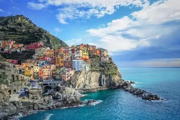 A village of colorful houses built into a rocky cliffside overlooking a turquoise sea under a blue sky with white clouds.