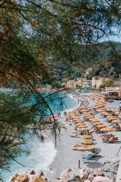 A crowded pebble beach viewed from above, filled with people relaxing under orange umbrellas next to turquoise water.